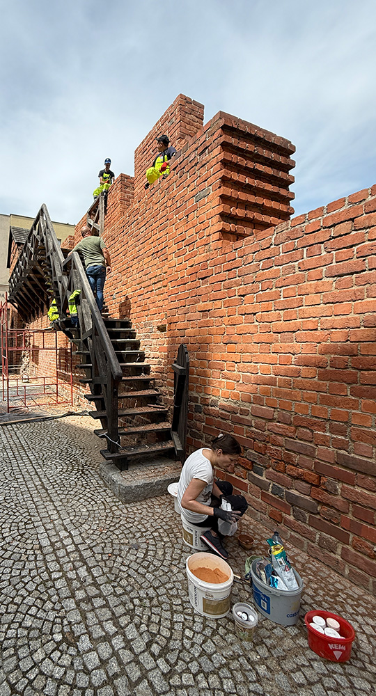 Prace konserwatorskie przy ceglanej zabytkowej ścianie, z osobą wykonującą uzupełnienia zaprawy przy murze i ekipą pracującą na drewnianych schodach oraz koronie muru.