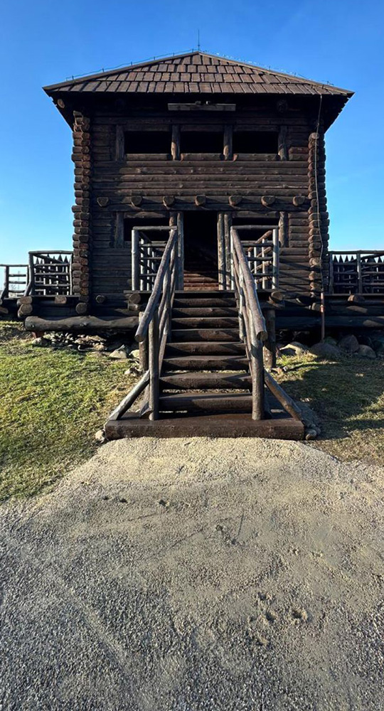 Drewniana strażnica zbudowana z masywnych bali, z prowadzącymi do wejścia schodami i barierkami, stojąca na otwartej przestrzeni pod czystym niebem.