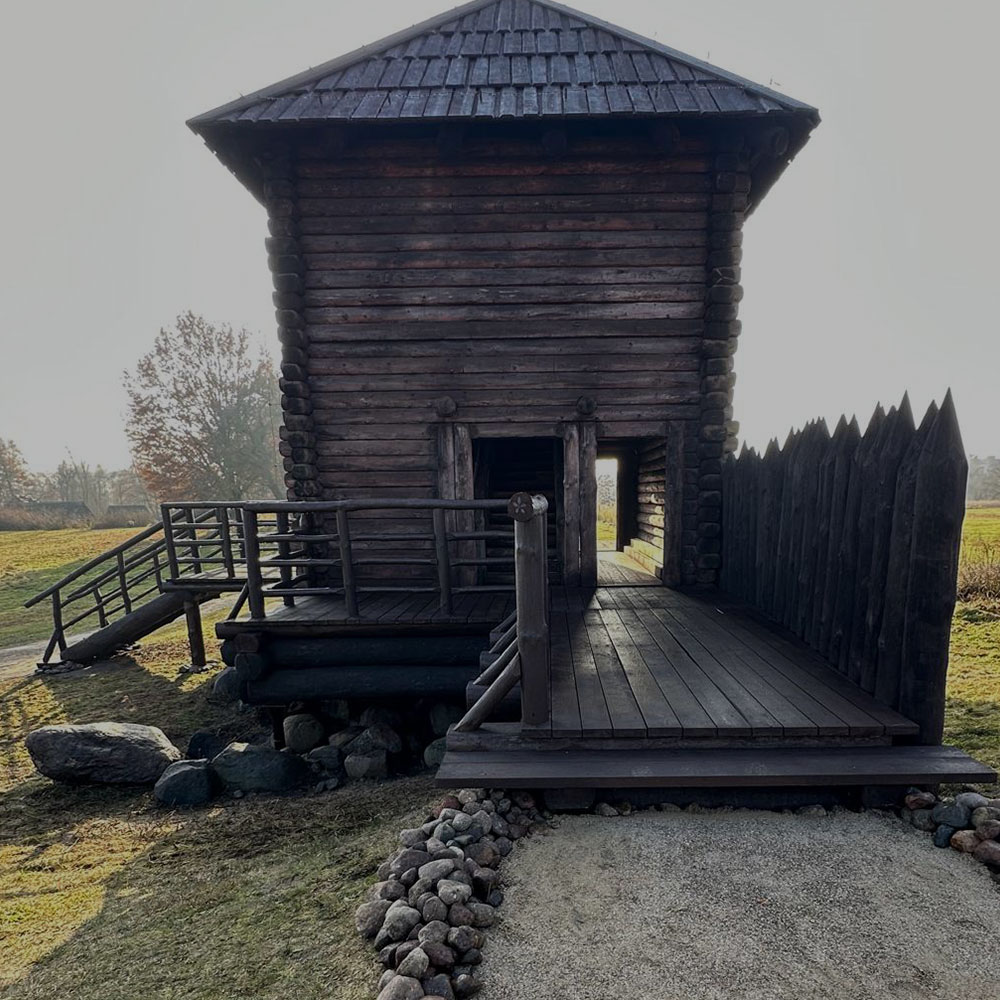 Drewniana historyczna wieża z ciemnych bali z rampą i pomostem, otoczona palisadą z zaostrzonych pali. Konstrukcja stoi na otwartej przestrzeni wśród traw i kamieni.