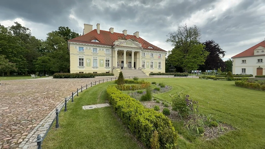 Okazały pałac o jasnej elewacji i czerwonym dachu z reprezentacyjnym portykiem wspartym na kolumnach, otoczony zadbanym ogrodem z nisko strzyżonymi żywopłotami i trawnikiem.