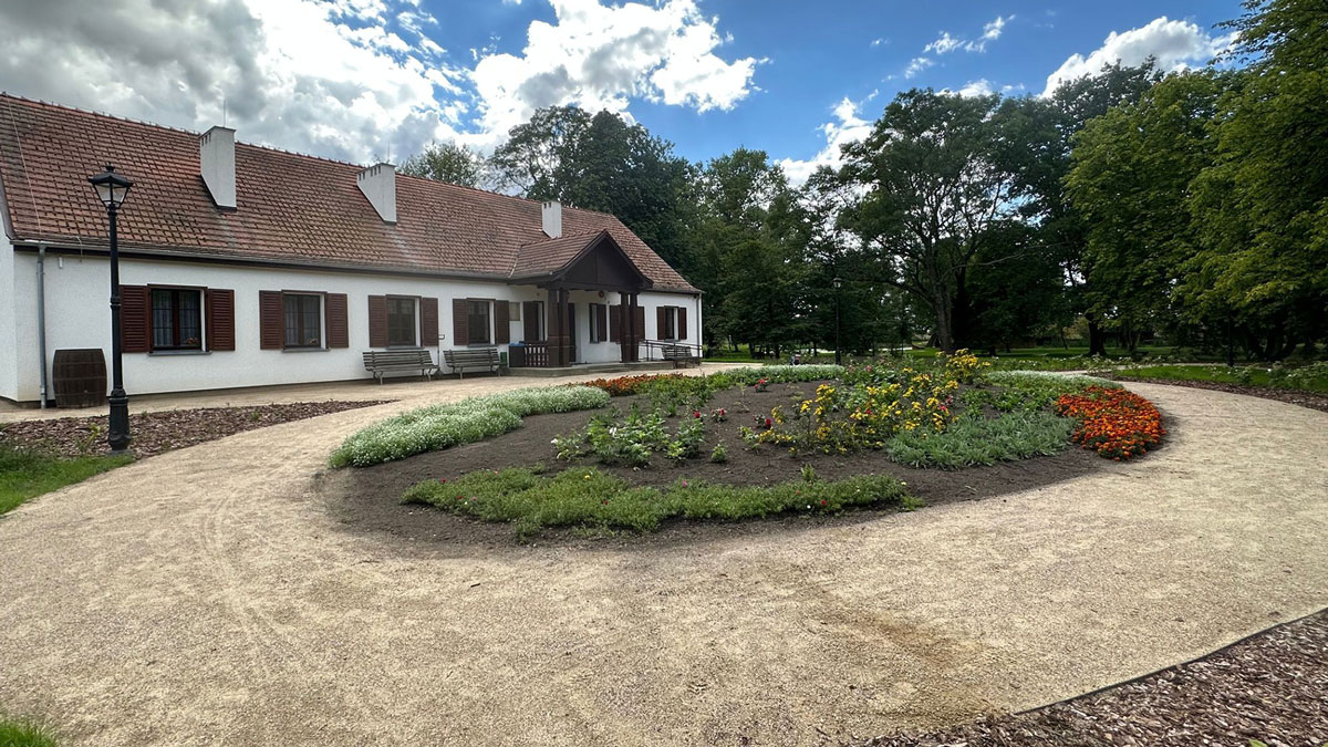 Parterowy biały dwór z drewnianymi okiennicami i werandą, stojący przy żwirowej ścieżce otaczającej kolorową rabatę kwiatową. Wokół rosną duże drzewa i rozciąga się parkowa zieleń.