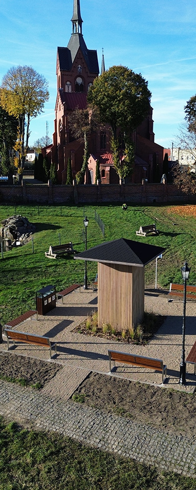 Nowoczesna drewniana tężnia stojąca na placu z żwirową nawierzchnią, otoczona ławkami i stylizowanymi latarniami. W tle widać zielony teren oraz ceglaną świątynię z wysoką wieżą.